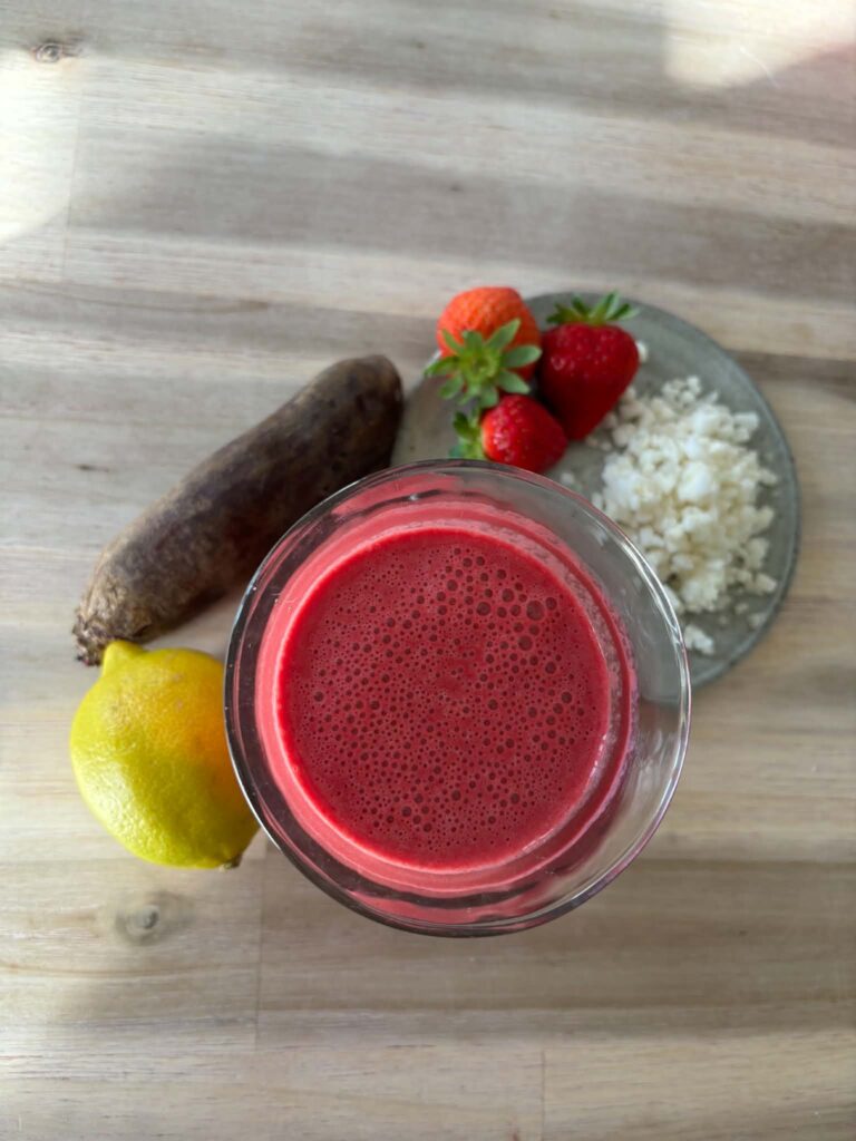 Glas med rødbedesmoothie omgivet af rødbede, citron, jordbær og blomkålsris på et træbord.